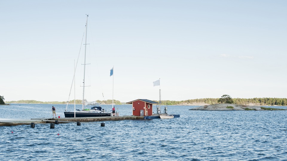 Segelbåt med blått skrov ligger längsmed brygga. Husarö Handel & Sjömack i Österåkers skärgård. 