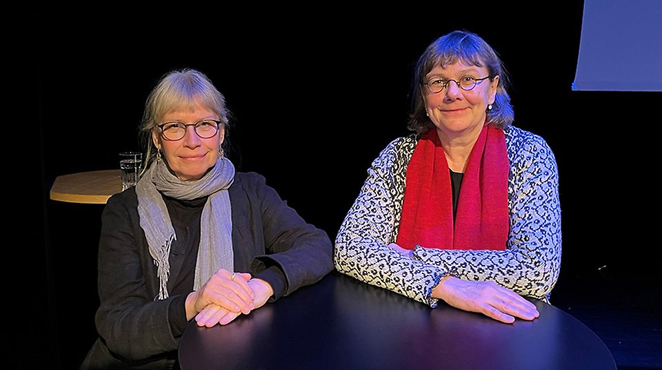 Två damer sittandes bakom ett svart bord ler in i kameran.