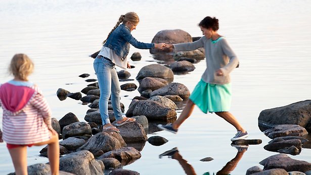 Tre tjejer går på några stenar som syns ovan vattenytan. 