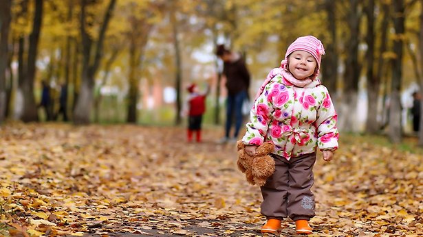 Ett barn går på en stig som är fyll med höstfärgade löv. I bakgrunden syns en vuxen och ytterligare ett barn.