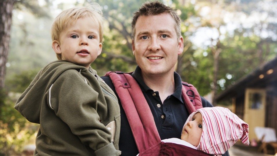 En föräldraledig pappa med sin bebis i en sele håller ett större barn på andra axeln. 