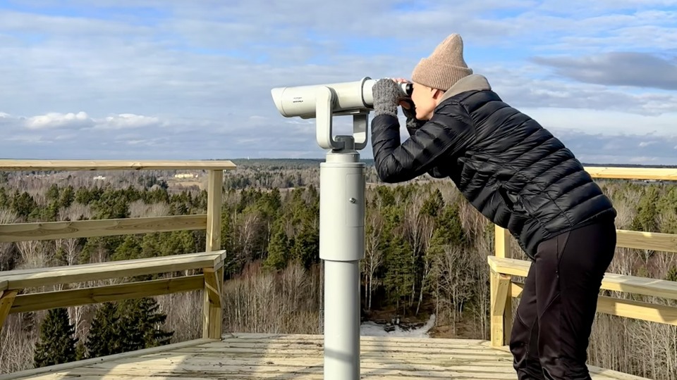 Fritidsklädd kvinna står på utsiktsplats i trä och spanar i en fast kikare.