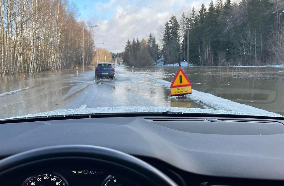 Foto taget inifrån en bil. Du ser en översvämmad vägsträcka och en varningsskylt vid sidan av vägen.