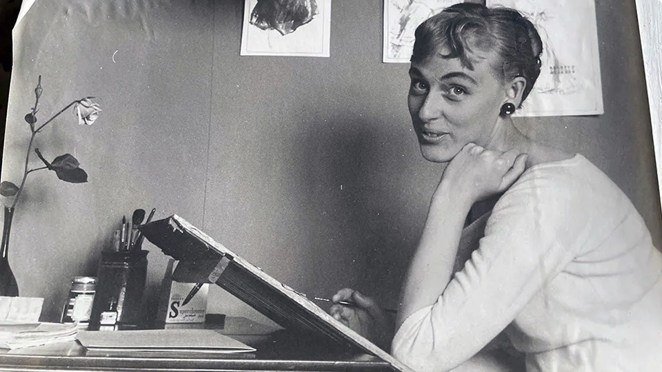 Ett svartvitt foto från 1950-60 talet. En kvinna sitter framför ett bord med ett litet staffli framför sig.