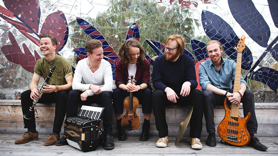 En grupp på fem personer sitter utomhus framför en mosaikvägg. De skrattar och har musikinstrument i händerna.