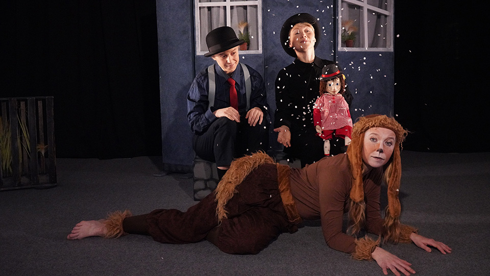 Tre skådespelare klädda för sina roller. Två har plommonstop och hängslen. En ser ut som en hund.