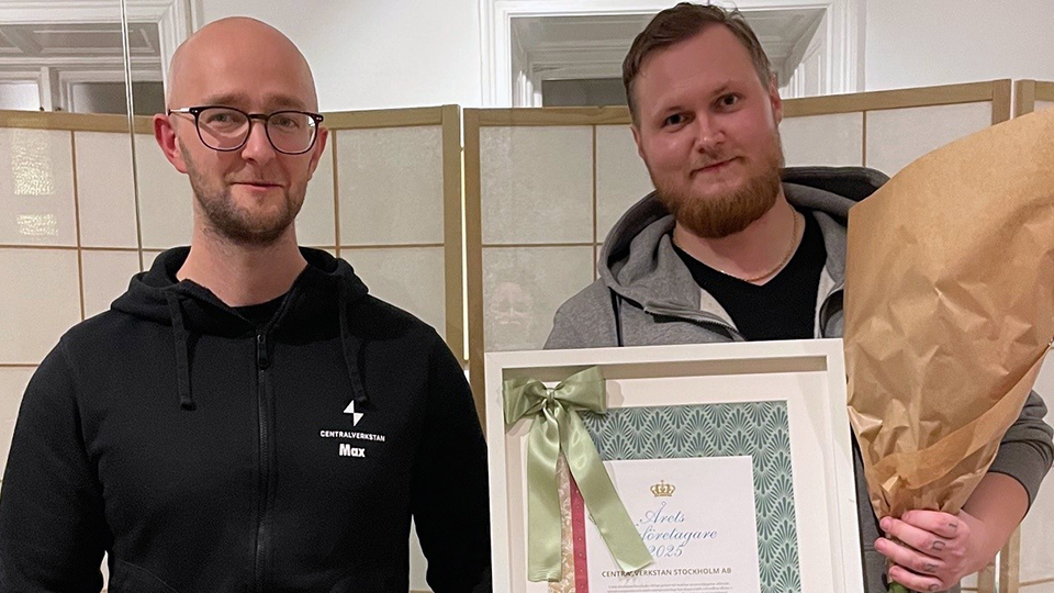 Två män ler in i kameran. De håller i ett diplom och ser glada ut.