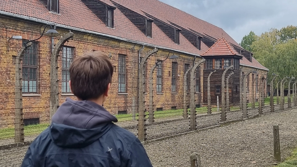 Person i mörkblå jacka med luva syns bakifrån. I bakgrunden syns en brun tegelbyggnad. Himlen är grå och gräset är grönt