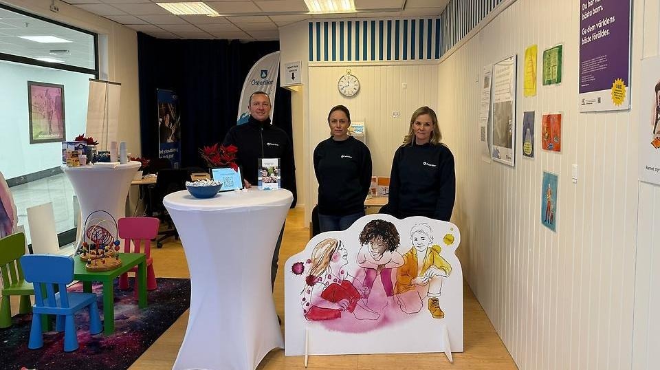 Tre medarbetare från Österåkers kommun står i en bemannad informationshörna med ståbord, barnhörna och färgglada affischer om föräldraskap, framför en stor skylt med illustrerade barn.