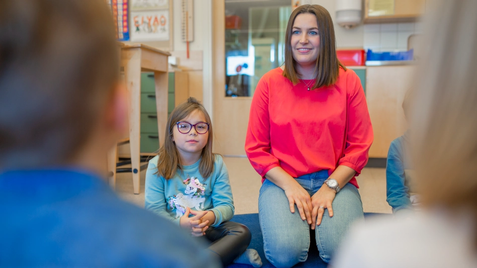 Bilden visar en skolmiljö inomhus. På golvet sitter barn och en pedagog i en ring.