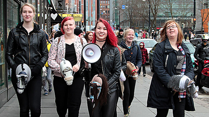 Unga kvinnor promenerar i stadsmiljö och de har sina käpphästar med sig. En av kvinnorna har en megafon i handen.