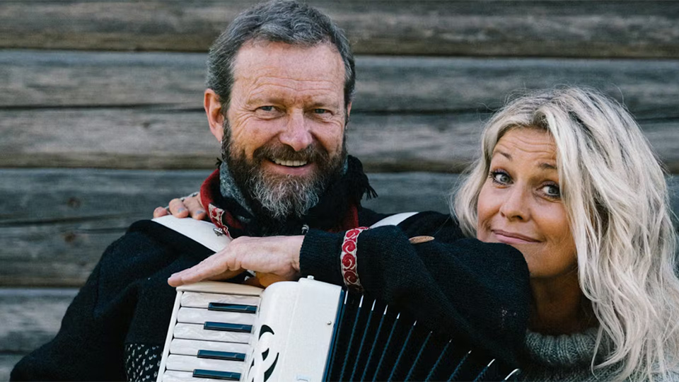 En man och en kvinna står bredvid varandra och ler in i kameran. Mannen har dragspel och grått skägg; kvinnan har långt, blont hår.