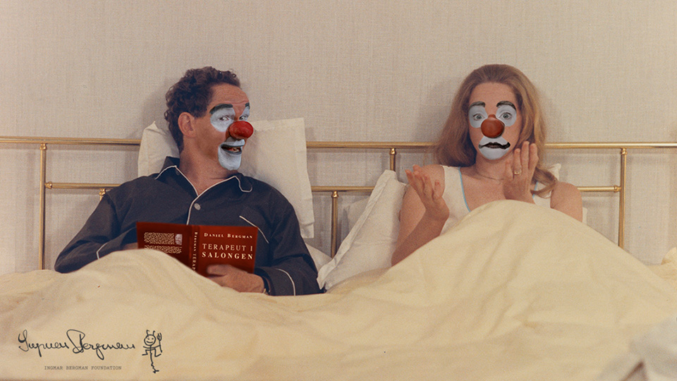 En man och en kvinna ligger bredvid varandra i en dubbelsäng. De är sminkade som clowner. Mannen läser boken "Terapeut i salongen".