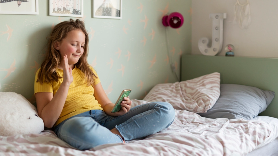 En barn sitter i en säng i sitt rum. Barnet ser glatt ut och har en mobiltelefon i handen.