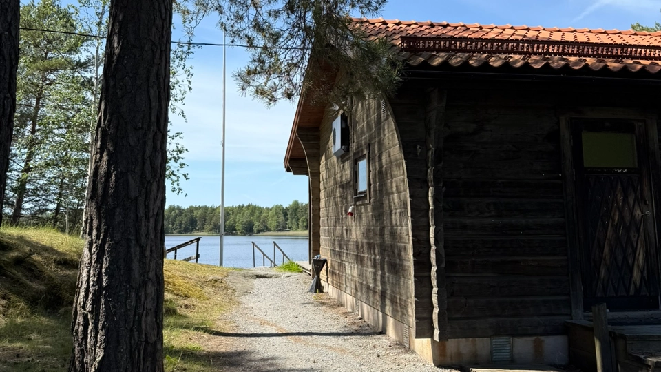 Bilden föreställer ett svart hus. Bredvid huset finns några tallar. I bakgrunden finns en sjö.