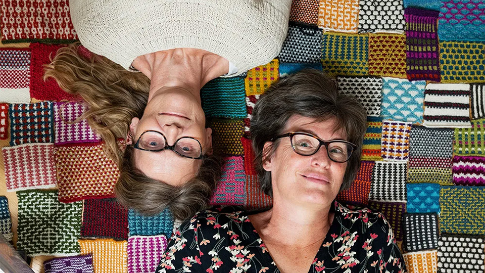 Två kvinnor ligger på ett stickat lapptäcke. De är fotade ovanifrån och ligger skavfötters.