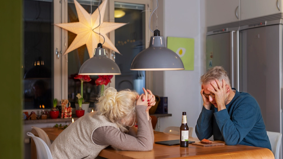 Ett par sitter vid ett matbord. De ser uppgivna ut. Köket är dekorerat med julpynt. I fönstret hänger en julstjärna. På bordet står en ölflaska.