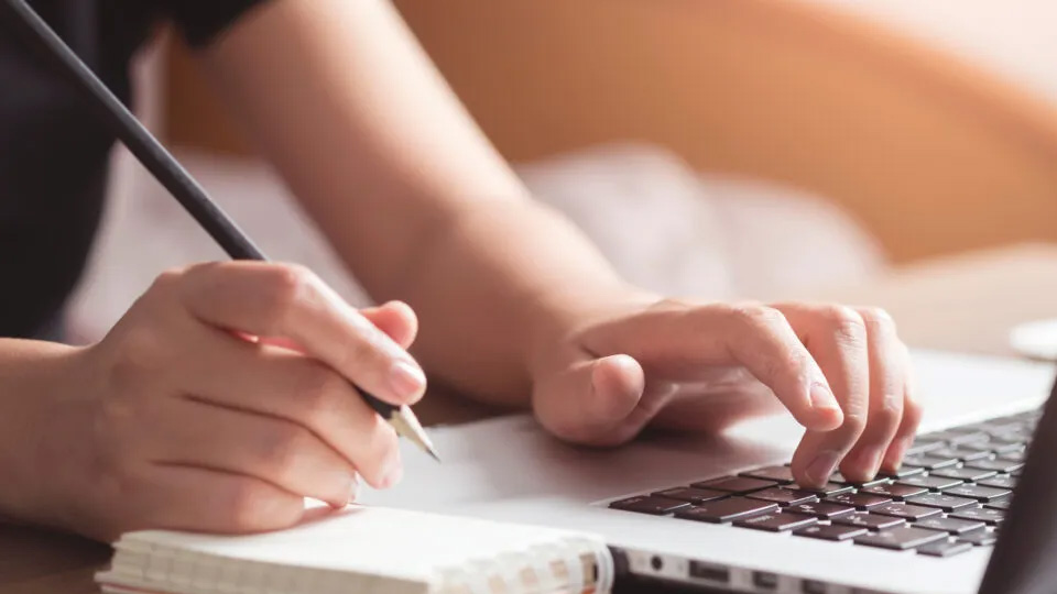 Vi ser en persons händer. Högerhanden håller en blyertspenna över ett block och vänsterhanden skriver på ett tangentbord.