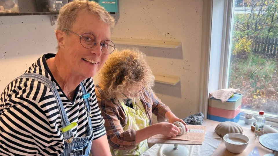 En kvinna i snickarbyxor ler in i kameran. Vid ett bord med lera sitter en person och skapar ett fantasidjur.