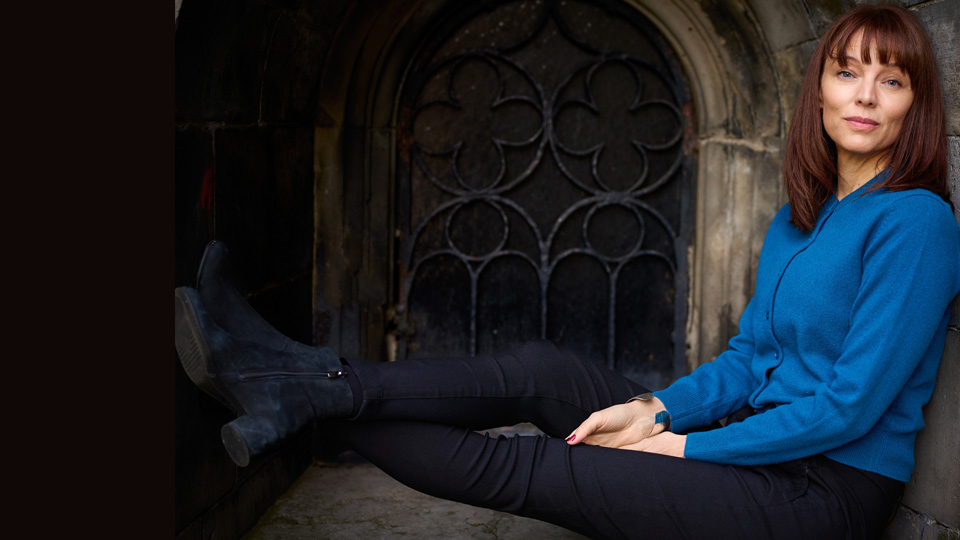 En kvinna med lugg och lång page sitter under ett stenvalv.