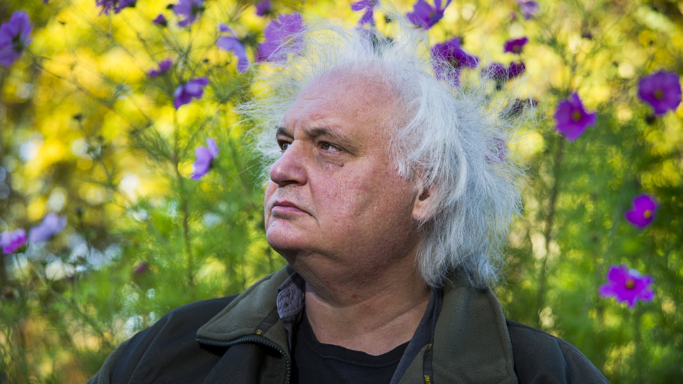 En äldre man med stort, vitt hår i rufsiga lockar. Han står utomhus framför blommande ängsblommor.