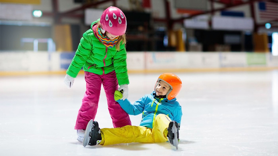 Två barn åker skridsko i en inomhushall. En av dem har ramlat och får hjälp av den andre att ta sig upp.
