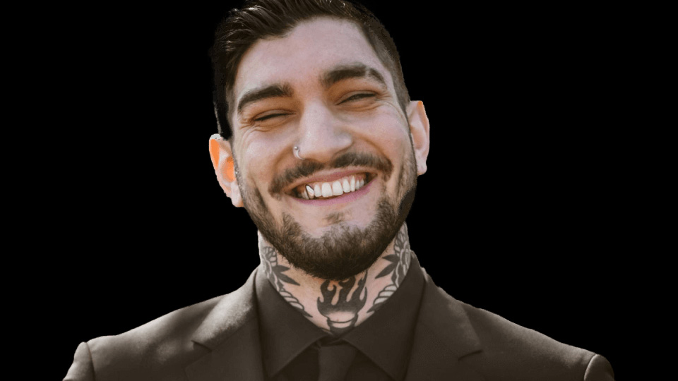 En ung man med brett leende och tatueringar på halsen. Han har guldtand, skjorta och kavaj.