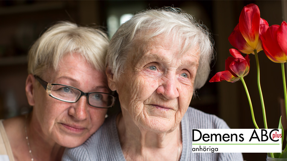 En äldre kvinna sitter bredvid en blommande krukväxt. Bredvid henne, med huvudet lutat mot den äldre kvinnans axel sitter en yngre. Möjligen hennes dotter.