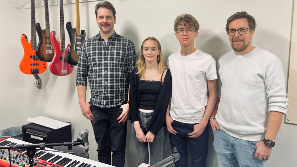 På fotot syns två lärare och två elever stå bredvid gitarrer och basar som hänger på väggen och två keyboards framför. 