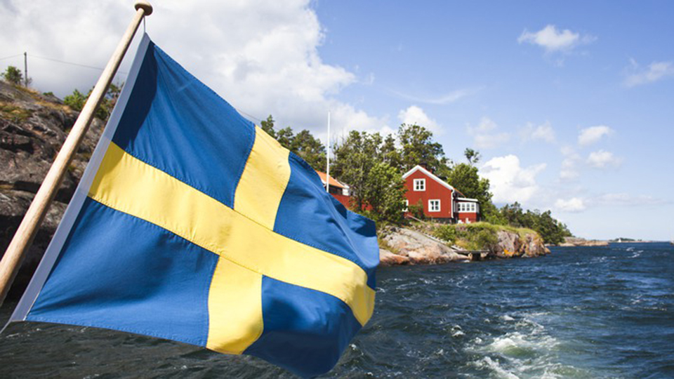 En svensk flagga vajar från aktern på en båt. I bakgrunden ses en röd stuga.