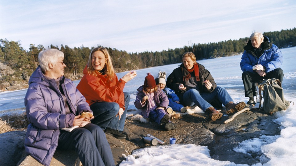 Bilden visar flera personer i blandade åldrar som sitter ner tillsammans och fikar i ett soligt vinterlandskap 