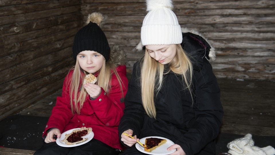 Bilden visar en tonåring och ett yngre barn som sitter i ett vindskydd. De har vinterkläder och äter våfflor