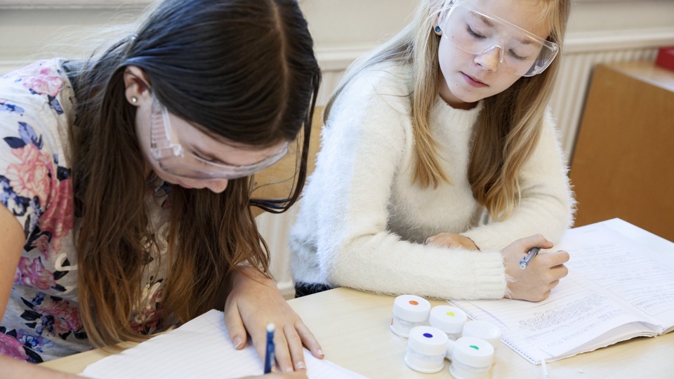 Två tjejer sitter i skolmiljö med skyddsglasögon och utför ett experiment.