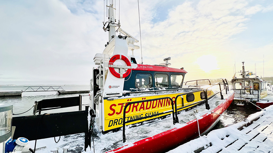Bilden visar en motorbåt med texten Sjöräddning  och namanet Drottnings Silvia. Båten är vit med rött skrov och har en livboj hängandes på sidan. Framför båten skymtar en annan båt. Det ligger snö på bryggan.