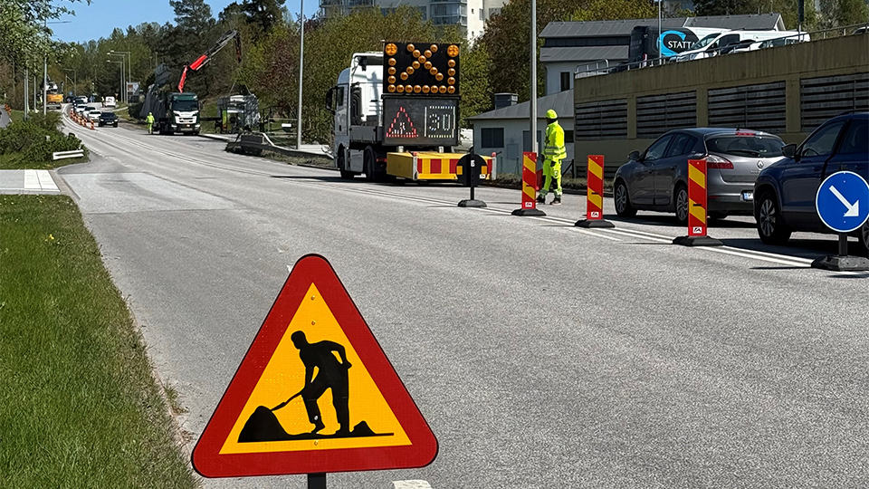 En trafikskylt som varnar för vägarbete på en väg. En trafikvakt syns i bakgrunden och man ser att arbete pågår på vägen.