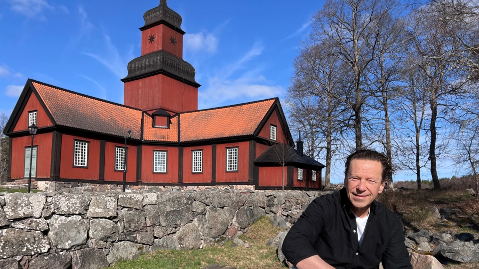 Porträttbild av Roger Persson sitter framför en faluröd kyrka i trä. Solen skiner och himlen är blå.