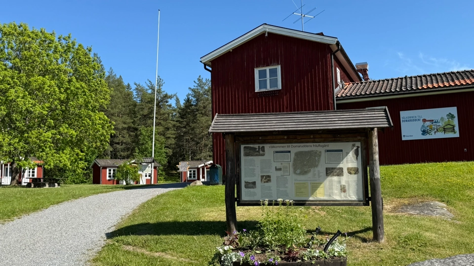 Faluröd byggnad i friluftsmiljö. Byggnaden har en skylt där det står Välkommen till Domarudden.