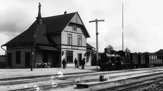 Stillbild ur Roger Perssons film om järnvägens ankomst till Österåker. Stationsbyggnad med några personer syns.