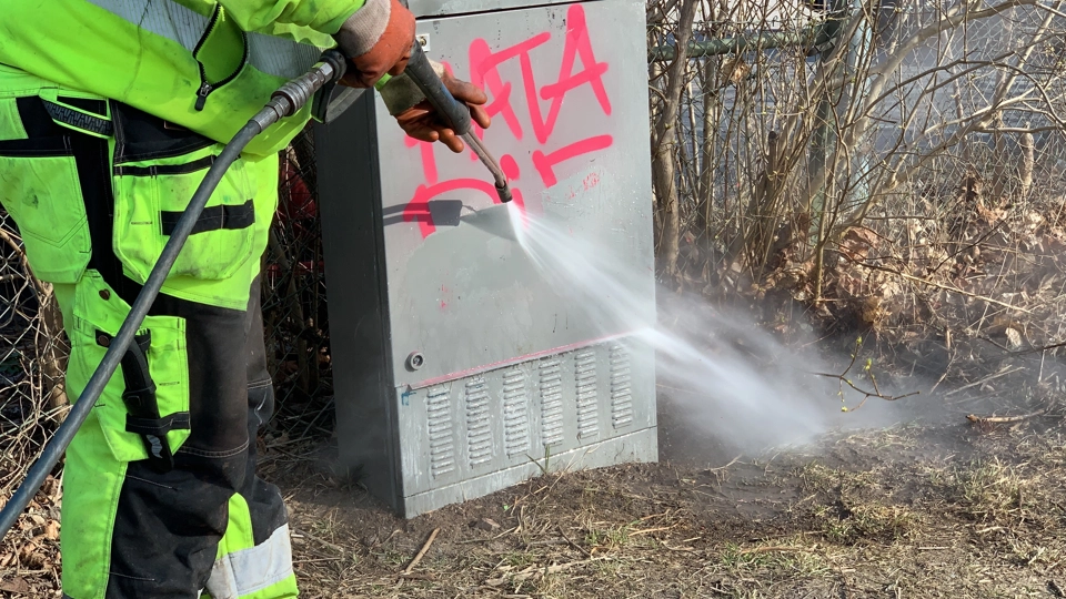 du ser en person i arbetskläder som tar bort klotter på ett elskåp med en högtrycksspruta.