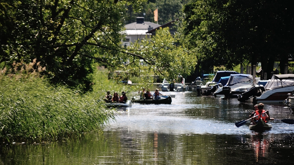 Bilden visar Åkers kanal. Träden är grönskande och i kanalen paddlar flera kajaker.