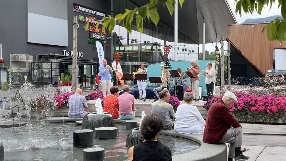 Flera musiker med blåsinstrument spelar utomhus vid Åkersberga centrum under Seniordagarna 2024. Publik sitter vid fontänen framför scenen.