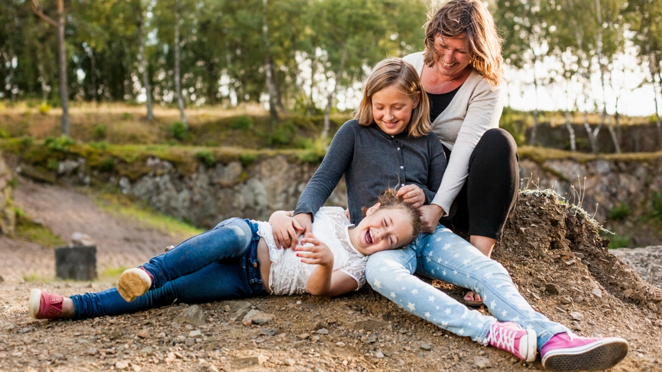 En familj med en mamma och två döttrar sitter på marken utomhus. Ett av barnen ligger i mammans knä. De ser väldigt glada ut.