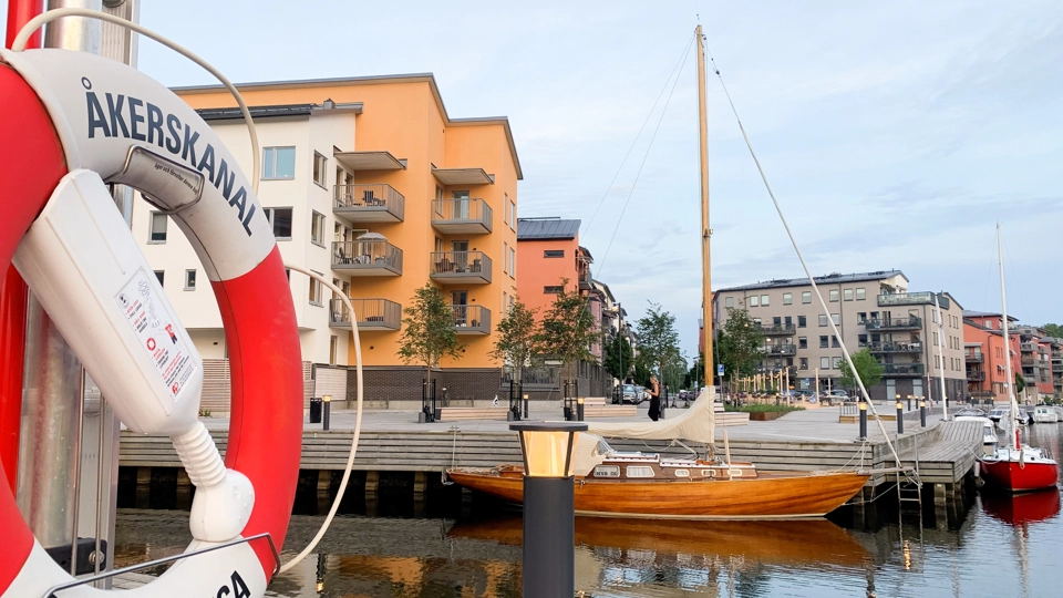Ett segelfartyg i trä förtöjd vid en gästbrygga i en kanal omgiven av flerfamiljshus och bryggor en solig sommardag.