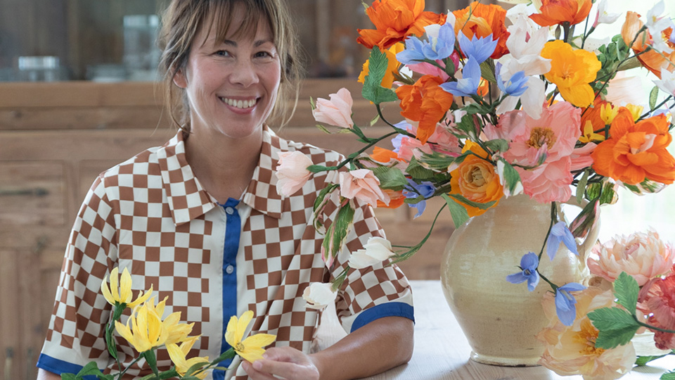 En leende kvinna sitter bredvid en stor bukett med färgglada pappersblommor.