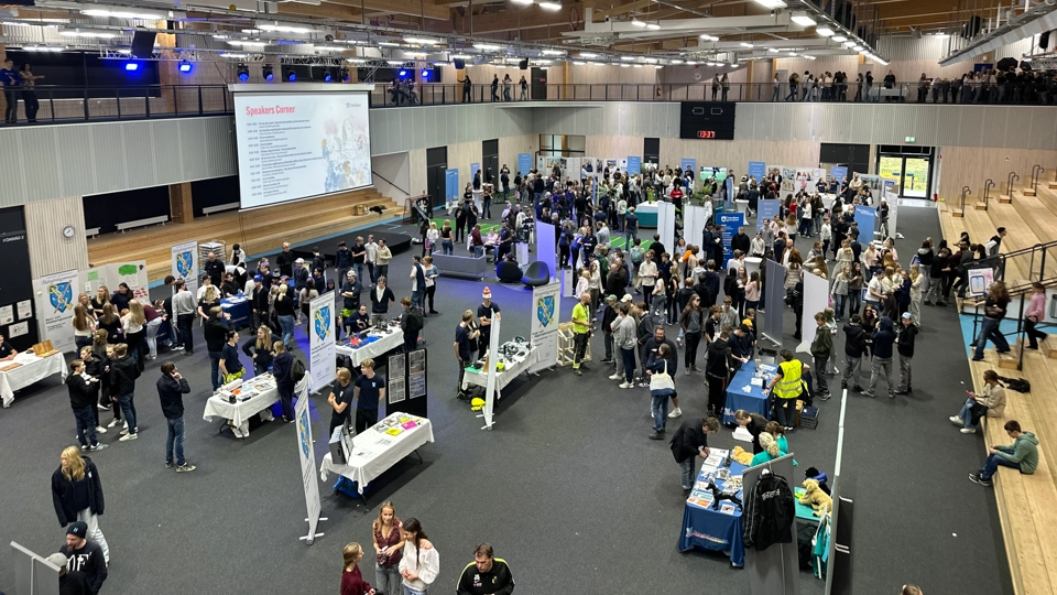 Bilden visar en stor idrottshall inifrån. På golvet står flera utställare. Det är mycket folk.