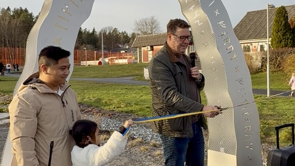 En person klipper ett blågult band. Han står i en båge som är ett konstverk med runskrift. Ett barn håller i bandet på ena sidan.