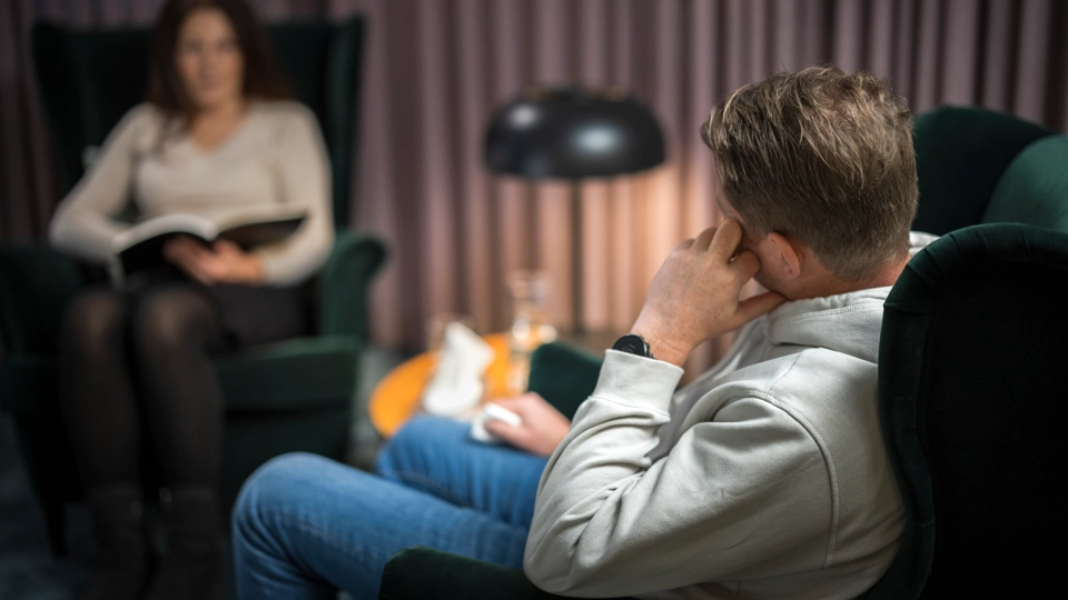 En man och en kvinna sitter mitt emot varandra. Mannen håller i en näsduk. Kvinnan håller i en anteckningsbok.