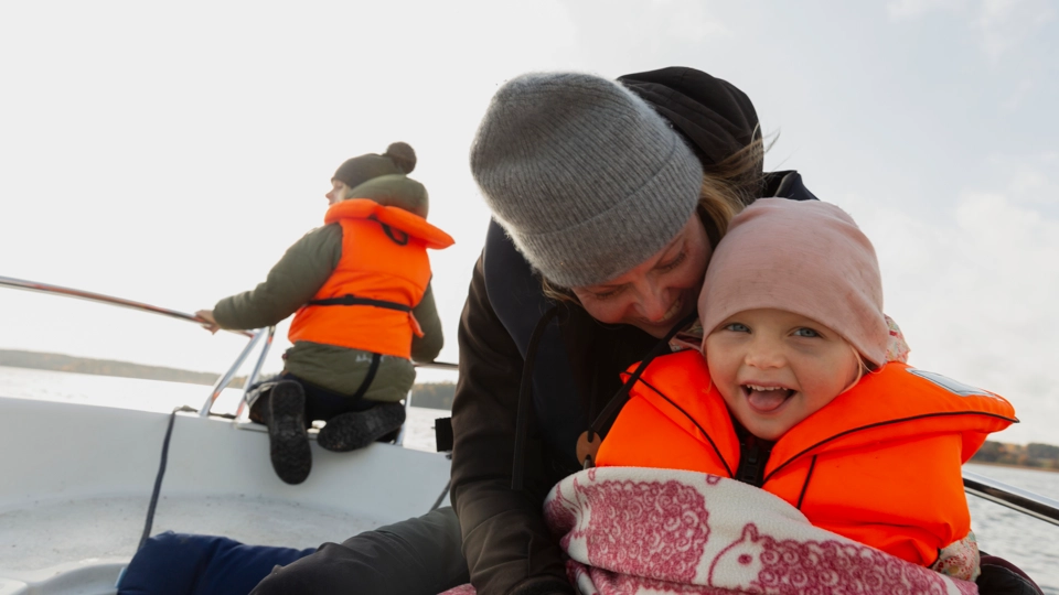 En förälder med ett barn sitter vinterklädda i en båt. De har flytvästar på och ser glada ut.