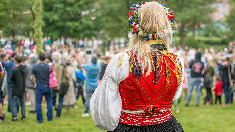 En gräsplan en solig sommardag. En stor grupp människor samlade kring en midsommarstång. Nära kameran står en kvinna vi ser bakifrån. Hon är klädd i folkdräkt och har blomsterkrans i håret.