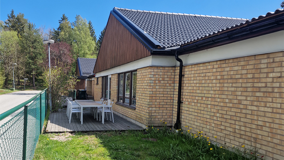 En tegelbyggnad med mörkt tak och trädetaljer på fasaden. Framför huset finns en liten altan med ett vitt bord och fyra stolar. Intill altanen går ett grönt staket längs en trottoar, och i bakgrunden syns grönskande träd samt en gata med lyktstolpar. Himlen är blå och vädret ser soligt ut.